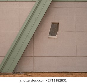 Geometric architecture clash: A sharp diagonal green steel beam cuts across a plain beige concrete wall featuring a single, irregularly shaped window.