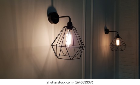 Geometric pendant light closeup on blurry background. Diamond shape wire lampshade with light bulb inside. Sconce lamps hanging on wall background with shadows and copy space. Interior lighting.