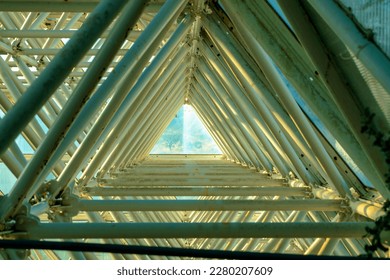 Geometric tunnel with triangular metal beams and scaffolding in greenhouse building or structure with glass. View to end of area with light outside at the end with dark shadow bars.
