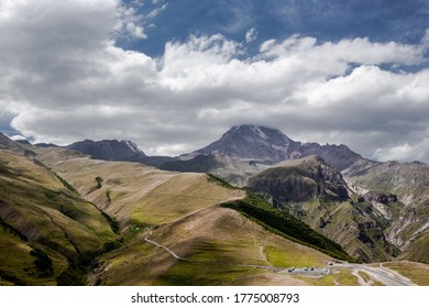 Georgia.  Caucasus. Alpine Road, Mountain Fault, Green Meadows & Rock Hills On Background Of Mount Kazbek Wrapped By White Clouds On Blue Sky