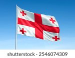 The Georgian flag with red crosses on a white background waves against a clear blue sky. The flag of Georgia features a large cross and four smaller crosses. National flag of Georgia.