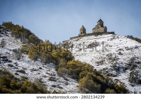 Gergeti Trinity Church (Tsminda Sameba Church) stands prominently atop a snow-dusted mountain slope under a clear blue sky in Stepantsminda, Georgia. The historic stone church is surrounded by sparse autumn foliage displaying shades of yellow and green, highlighting the remote and serene mountain landscape.