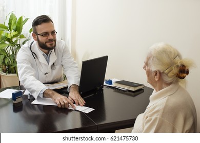 Geriatrician Doctor With A Patient In His Office. Old Woman At The Doctor Geriatrician. A Doctor At Work. He Shows A Finger And Translates The Patient Record On The Prescription.