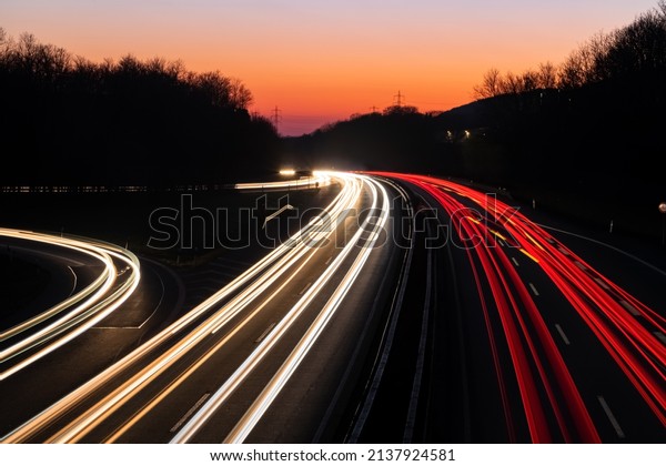 German Autobahn A46 at dusk on a winter evening\
in Iserlohn Sauerland at junction driveway town centre, long time\
exposure with light traces of passing car lights. Orange sky\
gradient after sunset.