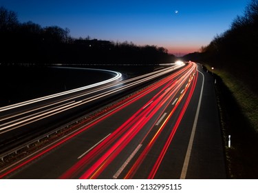 German Autobahn A46 At Evening Twilight In Winter In Iserlohn Sauerland At Junction Driveway Town Centre, Long Time Exposure With Light Traces Of Passing Car Lights. Blue Sky Gradient With Moon.