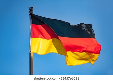 German flag in black-red-gold color on a sunny day in Germany. National yymbol of the “Bundesrepublik Deutschland“. Flag pole and wavy fabric blown in the wind on a blue sky day. Intense sunlit colors