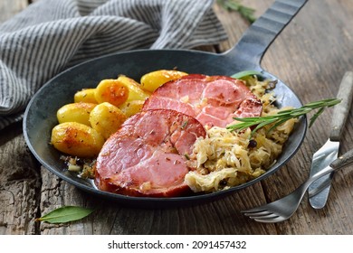Deutsches Essen - Geräucherter Schweinehals mit Sauerkraut und gebratene Babykartoffeln in einer eisernen Pfanne auf einem alten Holztisch