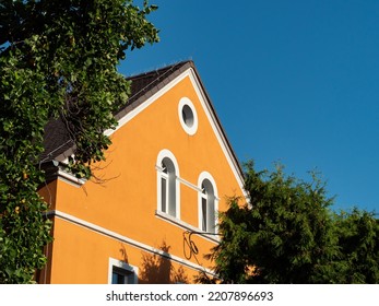 An German house facade in between green tree crowns. Old building exterior with a gable roof in front of a blue sky. Ancient architecture in East Germany in a rural area. Renovated with orange color.