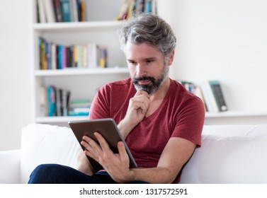 German Middle Aged Man With Grey Hair Reading E-book With Digital Tablet Indoors At Home