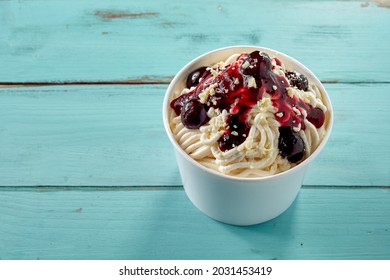 German Vanilla Spaghetti Ice-cream Sundae With Black Cherries And Fruity Sauce In A Takeaway Tube Viewed Top Down Over Blue Wood Table With Copyspace For Menu Advertising