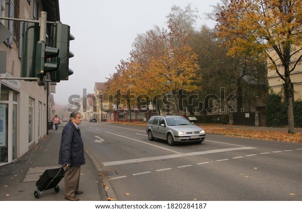 Germany 2007.10.10 an Elderly man with a bag\
on wheels stands at a pedestrian crossing with a traffic light on a\
cloudy autumn day. City\
life.