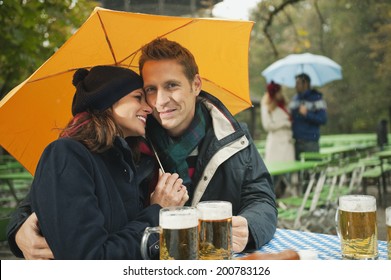 Germany, Bavaria, English Garden, Couple Sitting Under Umbrella In Beer Garden Raining