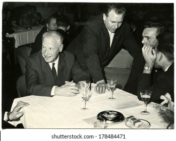 GERMANY - CIRCA 1950s: An Antique Photo Of Group Of Businessmen At A Table In The Restaurant, They Are Faced With Alcohol Glasses, Ashtray, Lies A Pack Of Cigarettes