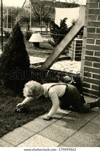 GERMANY - CIRCA 1960s: An antique\
photo of little boy toy car rolls by law, kneeling on the\
path