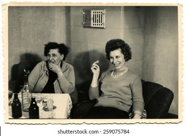 GERMANY - CIRCA 1960s: Two Smiling Women Sitting At The Banquet Table In Front Of Them Is A Bottle Of Vodka And Two Bottles Of Beer, A Young Woman Smoking A Cigarette