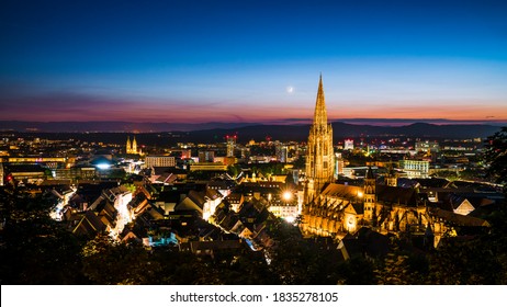 Germany, Freiburg im Breisgau, Magical blue hour atmoshpere of the illuminated skyline and minster cathedral, aerial view above with moon