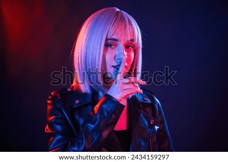 Gesture of be quiet, finger is by the lips. Woman with white hair is in studio with neon colors.
