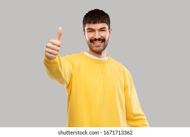 gesture and people concept - smiling young man in yellow sweatshirt showing thumbs up over grey background