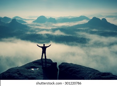 Gesture of triumph. Happy hiker in black.  Tall man on the peak of sandstone rock in national park Saxony Switzerland watching down to landscape.