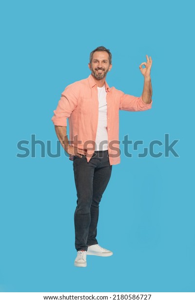 Gesturing OK handsome middle aged grey\
haired man happy smiling looking at camera wearing peach shirt\
isolated on blue background. Fit mature man saying im OK.\

