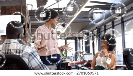 Gesturing presenter in pink blouse and dark pants speaking in open office, with monitors, UI icons. Collaboration, teamwork, brainstorming, strategy, modern, industrial, data