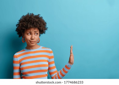Get away from me. Displeased curly woman pulls hand aside in stop or no gesture, being bothered by someone, denies problems, ignores something, dressed in casual striped jumper, isolated on blue wall - Shutterstock ID 1774686233