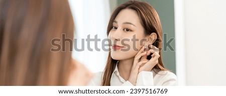 Get dress, asian young woman, businesswoman standing, hand putting earring or jewelry, wearing white shirt formal getting dressed getting ready before go to work looking reflection the mirror at home.