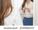 Get dress, beautiful asian young woman, businesswoman standing buttoning white shirt formal getting dressed, ready before go to work looking reflection the mirror in bedroom in the morning at home.