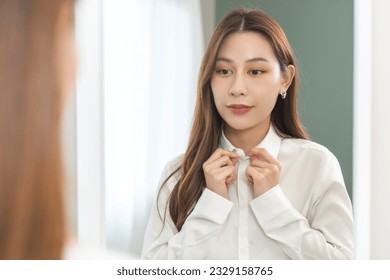 Get dress, beautiful asian young woman, businesswoman standing buttoning white shirt formal getting dressed, ready before go to work looking reflection the mirror in bedroom in the morning at home.