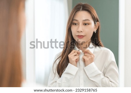 Get dress, beautiful asian young woman, businesswoman standing buttoning white shirt formal getting dressed, ready before go to work looking reflection the mirror in bedroom in the morning at home.