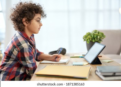 Get An Education. Enthusiastic Latin School Boy Wearing Glasses Making Notes, Having Online Lesson Using Digital Tablet While Studying At Home