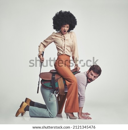 Get moving. A studio shot of an attractive woman in 70s wear riding a handsome man wearing a saddle while using a riding crop.