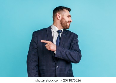 Get out! Portrait of angry man with beard wearing dark suit looking aside and pointing finger another way, asking to leave, dissatisfied expression. Indoor studio shot isolated on blue background.