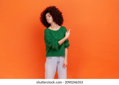 Get out! Woman with Afro hairstyle wearing green casual style sweater pointing away, scolding for bad result and showing exit, ordering to leave. Indoor studio shot isolated on orange background.