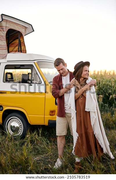 It\'s getting cold. Relaxed happy Hippie\
couple on trip in countryside. yellow retro style van vehicle\
camper traile in the background, man covers wife with blanket.\
camping, traveling,\
hitchhiking.