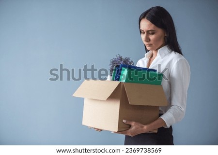 Getting fired. Beautiful young business woman in formal wear is holding a box with her stuff and looking sadly at it, on gray background