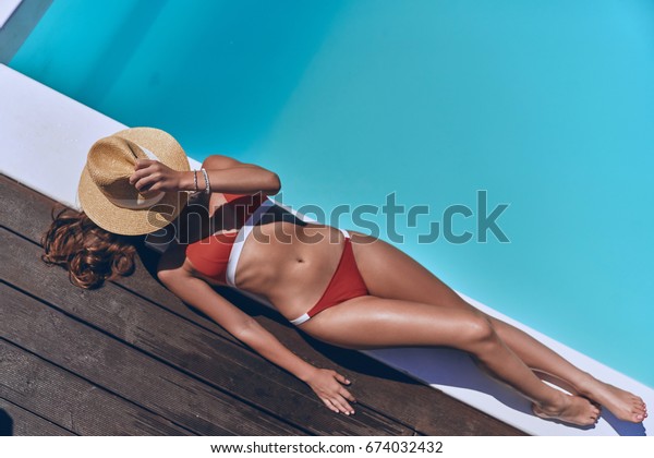 Getting golden tan. Full length top view of\
beautiful young woman in swimwear covering face with hat while\
sunbathing by the pool