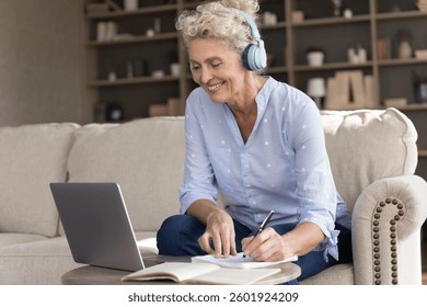 Getting knowledge in any age. Smiling senior woman in wireless headphones sit on sofa engaged in remote learning watch webinar training course via notebook handwrite notes in workbook after teacher