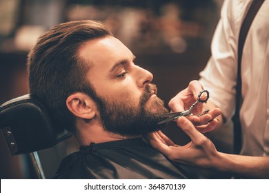 Getting perfect shape. Close-up side view of young bearded man getting beard haircut by hairdresser at barbershop - Shutterstock ID 364870139