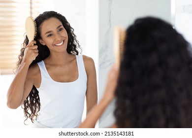 Getting Ready. Beautiful Arab Female Combing Her Curly Hair With Brush While Standing Near Mirror In Bathroom, Young Happy Woman Looking To Her Reflection And Smiling, Selective Focus With Free Space