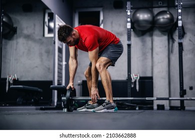 Getting ready for fitness exercise, preparing barbell weights. A shot of a handsome man in a red shirt and shorts setting up equipment for strong functional training in a modern sports gym - Shutterstock ID 2103662888