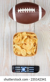 Getting Ready To Watch A Bowl Game. High Angle Shot Of An American Football, A Bowl Of Potato Chips And A TV Remote On A White Wood Table. Vertical Format