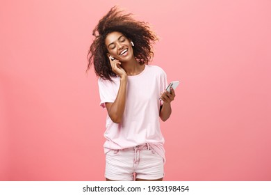 Getting rid of worried with good tunes in ears. Pleased attractive young african woman in trendy outfit listening music in wireless earphones touching earbud smiling dancing and holding smartphone