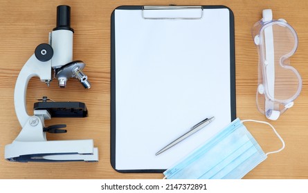 Getting Started On A New Research Project. High Angle Shot Of A Microscope, Clipboard, Pen, Protective Glasses And Face Mask On A Table.