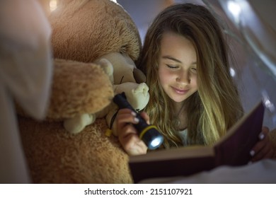 Getting Stuck In A Good Story. Cropped Shot Of A Young Girl Reading A Book By Torchlight Under A Blanket Fort At Home.