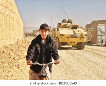 GHAZNI, AFGHANISTAN - NOVEMBER 2010: An Afghan Boy Rides His Bike As A Polish And US Soldiers From Task Force White Eagle Patrol His Village.