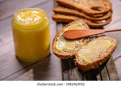 Ghee butter in glass jar with wooden spatula and sliced bread on wooden table.