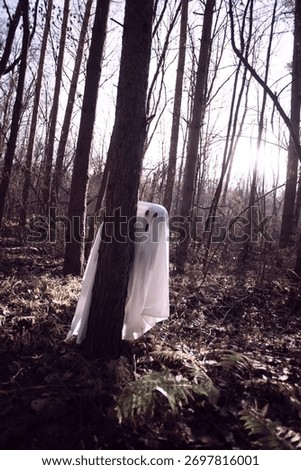 The ghost is covered with a white ghostly sheet in the autumn twilight forest. A creepy white ghost is peeking out from behind the trees