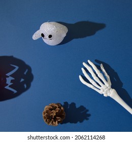 Ghost, Pine Cone And Hand Skeleton With Shadows, With A Shadow Of Halloween Pumpkin On A Blue Background. Flat Lay Concept.