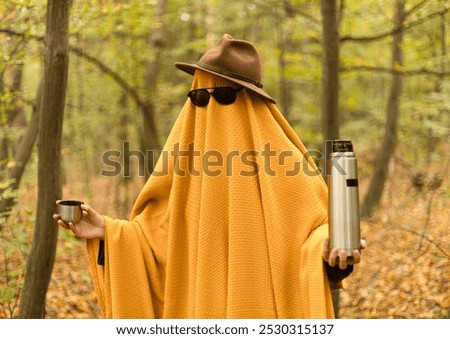 ghost in a yellow blanket, hat and glasses in the autumn forest with a thermos